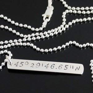 May include: A silver necklace with a silver bar pendant engraved with the coordinates "45° 29'14.6.65"N".