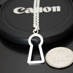 May include: A silver-colored keyhole pendant necklace. The pendant is a simple, open keyhole shape hanging from a delicate chain. A coin is next to the pendant for size comparison. The background is a black surface.
