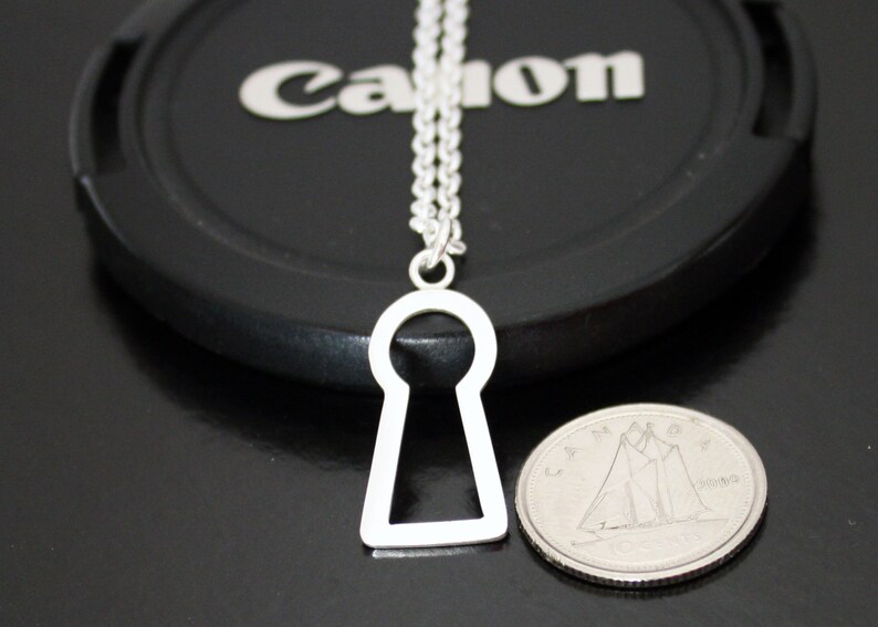 May include: A silver-colored keyhole pendant necklace. The pendant is a simple, open keyhole shape hanging from a delicate chain. A coin is next to the pendant for size comparison. The background is a black surface.