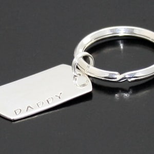 以下が含まれることがあります： 「DADDY」と刻まれた長方形のタグが付いたシルバーのキーホルダーです。