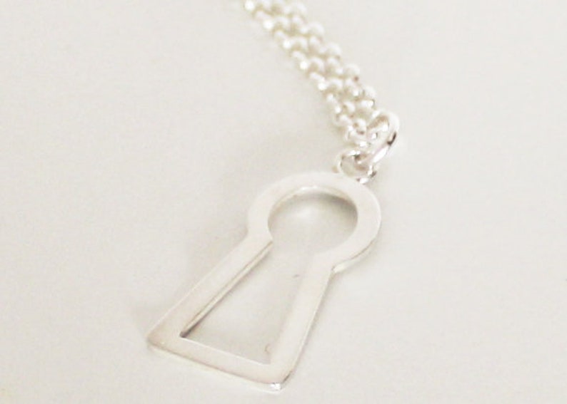 May include: A silver-colored keyhole pendant necklace. The pendant is a stylized keyhole shape with a rounded top and a rectangular base. The necklace chain is a delicate silver ball chain.