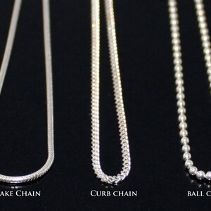 May include: Three silver chains, a snake chain, a curb chain, and a ball chain, are displayed against a black background. The chains are all the same length and are hanging straight down.