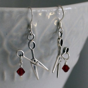 May include: A pair of silver earrings featuring a pair of scissors with a red crystal dangle on each earring.