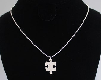 Puede incluir: Un collar de cadena de plata con un colgante de pieza de rompecabezas de plata. La pieza del rompecabezas tiene un pequeño corazón grabado.