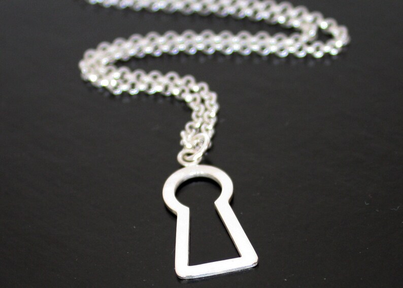 May include: A silver-colored necklace featuring a keyhole pendant. The pendant is a simple, open keyhole shape, and the chain is a delicate, linked design. The necklace is displayed against a dark background.