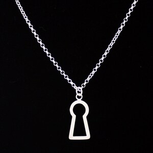 May include: A silver-colored necklace with a keyhole pendant. The pendant is a simple outline of a keyhole shape. The chain is a delicate link chain. The background is solid black.