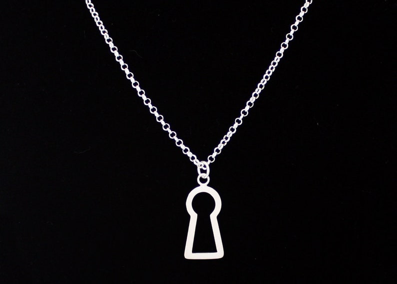 May include: A silver-colored necklace with a keyhole pendant. The pendant is a simple outline of a keyhole shape. The chain is a delicate link chain. The background is solid black.