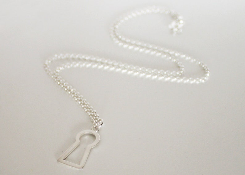 May include: A silver-colored necklace featuring a keyhole pendant. The pendant is a simple, geometric design with a rectangular shape and a circular opening. The chain is delicate and hangs gracefully.
