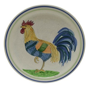 May include: A white ceramic plate with a blue, green, and yellow rooster design. The rooster is standing on a green patch of grass.