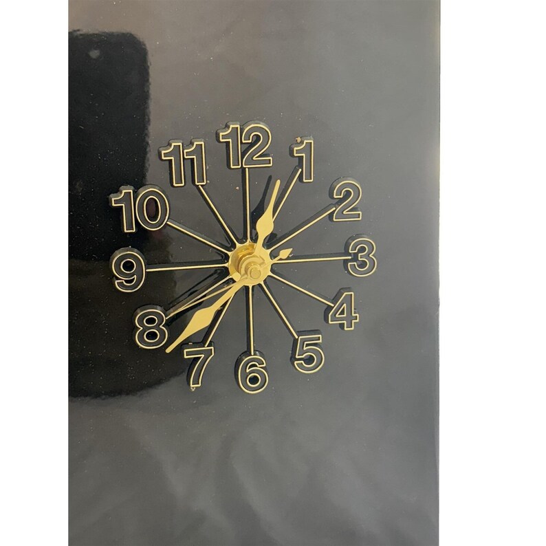 May include: A black wall clock with gold-colored hands and numerals. The clock face features a starburst design with the numbers 1 through 12. The hands are positioned to indicate the time.