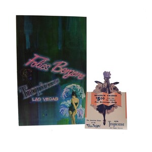 May include: Vintage Tropicana Las Vegas advertisement featuring the Folies Bergere show. The artwork includes a woman in a swimsuit, neon text, and a separate card with a minimum charge notice.