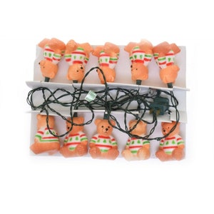 May include: A string of Christmas lights featuring 10 small, brown teddy bear figurines wearing green and red sweaters. The lights are packaged on a white card with a black wire.