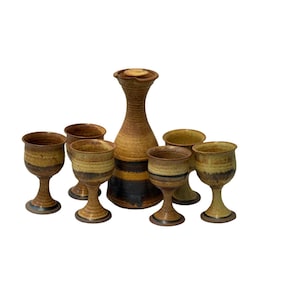 May include: A handmade ceramic drinkware set featuring a tall, slender pitcher with a lid and six matching stemmed goblets. The set has a rustic, earthy aesthetic with brown, tan, and black color bands.