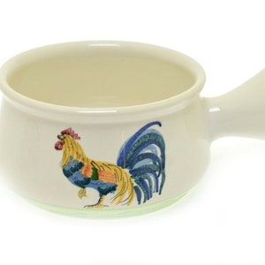 May include: A white ceramic soup bowl with a rooster design. The rooster is yellow, blue, green, and red. The bowl has a handle on the side.