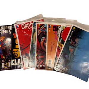 May include: A collection of comic books, each encased in a protective plastic sleeve. The covers feature vibrant illustrations of superheroes and comic book titles, including "Supergirl" and "Superman". The comics are stacked together, showcasing a variety of colors and designs.