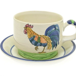 May include: A white ceramic teacup and saucer with a colorful rooster design. The rooster is painted in shades of blue, green, yellow, and red.