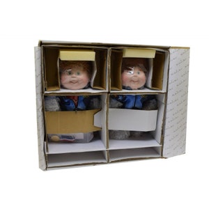 May include: Two Cabbage Patch Kids dolls in a cardboard box. The dolls are wearing blue and red jackets with a star design. The dolls are packaged in individual compartments with foam padding.