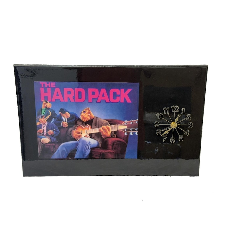 May include: Black rectangular clock with a picture of a band and the text "THE HARD PACK". The clock has a gold-colored clock face with black numbers and hands. The band is playing instruments.