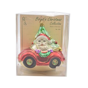 May include: A red glass ornament depicting Santa Claus driving a car with a Christmas tree on top. The ornament is part of the "Birgit's Christmas Collection" and is labeled "No. 103".