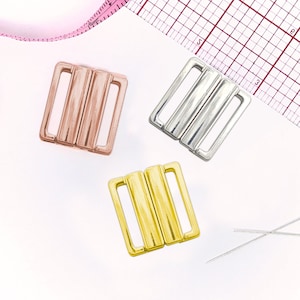 May include: Three metal bra clasps in gold, silver, and rose gold. The clasps are square shaped with three bars in the center.