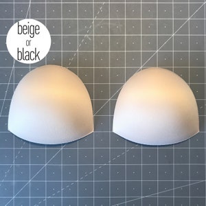 May include: Two beige or black bra cups, made of foam, on a gray grid background.