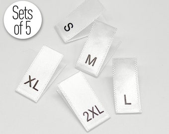May include: Five white satin clothing size labels with black text. The labels read "S", "M", "L", "XL", and "2XL".