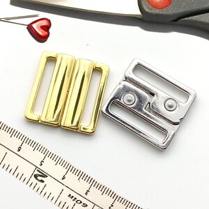May include: Two metal bra clasps, one gold and one silver. The clasps are square shaped and have a hook and eye closure.