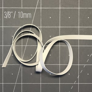 May include: A length of light gray elastic ribbon, 3/8 inch (9.5 mm) wide, is coiled on a dark gray cutting mat. The text "3/8" / 10mm" is in the upper left corner. The ribbon is flat and flexible.