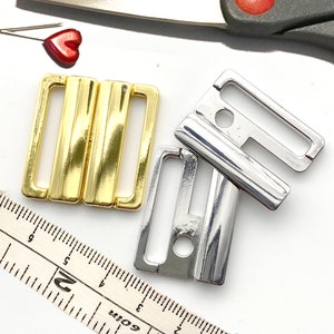 May include: Two bra clasps, one gold and one silver. The clasps are both rectangular with a small hole in the center. The gold clasp has a shiny finish, while the silver clasp has a matte finish.