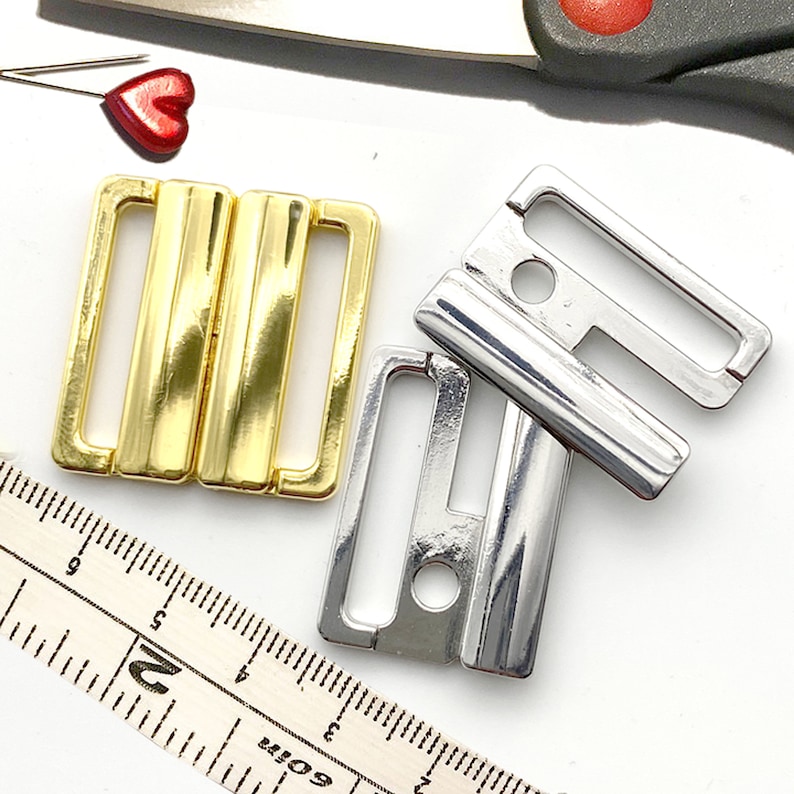 May include: Two bra clasps, one gold and one silver. The clasps are both rectangular with a small hole in the center. The gold clasp has a shiny finish, while the silver clasp has a matte finish.