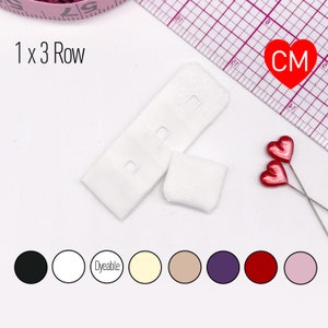 May include: White bra strap extender with three rows of hooks and eyes. The extender is 1 x 3 row. The extender is shown on a white surface with a ruler and a measuring tape. The extender is available in a variety of colors, including black, white, cream, beige, purple, red, and pink.