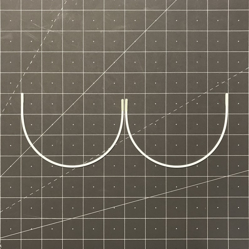 May include: Two white metal bra underwires, curved and joined at the centre, on a grey grid background.
