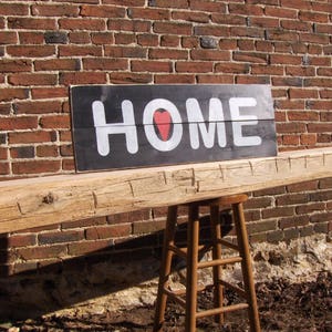 Puede incluir: Una viga de madera r&uacute;stica con un letrero negro que dice "HOME" con un coraz&oacute;n rojo en el medio de la palabra. El letrero est&aacute; montado en una pared de ladrillo.