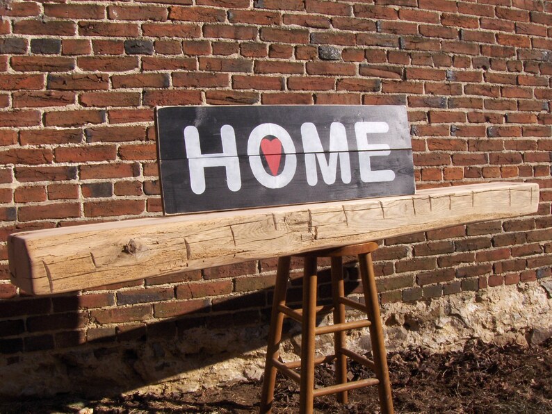 Puede incluir: Una viga de madera r&uacute;stica con un letrero negro que dice "HOME" con un coraz&oacute;n rojo en el medio de la palabra. El letrero est&aacute; montado en una pared de ladrillo.