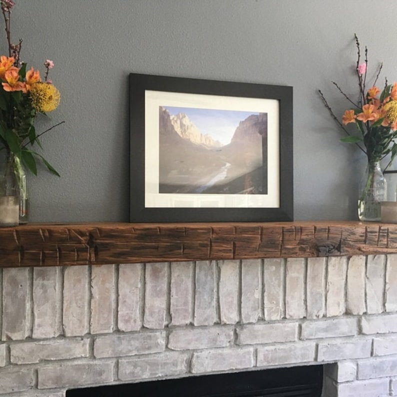 Puede incluir: Una repisa de chimenea de madera r&uacute;stica con un acabado marr&oacute;n oscuro se encuentra sobre una chimenea de ladrillo blanco. Una fotograf&iacute;a de paisaje enmarcada con una escena de monta&ntilde;a cuelga en la pared sobre la repisa. Dos jarrones con flores naranjas y rosas est&aacute;n sobre la repisa.
