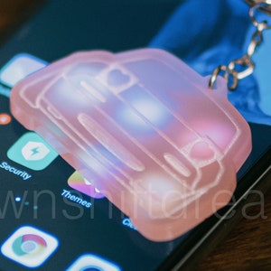 Peut inclure: Un porte-clés en forme de voiture rose avec un contour blanc et un anneau de clé argenté. Le porte-clés est posé sur un smartphone noir avec un écran bleu.