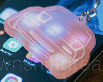 May include: A pink car-shaped keychain with a white outline and a silver key ring. The keychain is lying on a black smartphone with a blue screen.