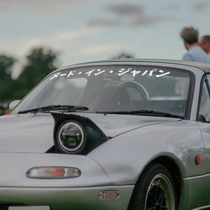 May include: A silver Mazda Miata convertible with black headlights and a white windshield banner that reads "オートイン・ジャパン". The car is parked on a grassy field.