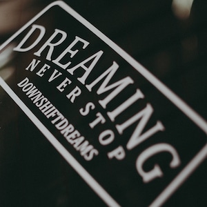 May include: A black and white sticker with the text "Dreaming Never Stop Downshiftdreams" on a black car window.