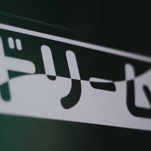 May include: White sticker with the Japanese word "Fly" in black lettering on a dark green surface.