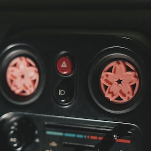 May include: Close-up of a car's dashboard with two pink cherry blossom air fresheners. The air fresheners are in the vents on either side of the dashboard. The dashboard is black and has a radio and climate control buttons.
