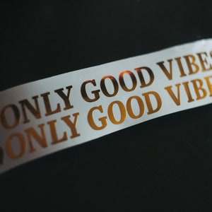 May include: White sticker with gold lettering that reads "ONLY GOOD VIBES" twice. The sticker has two small gold hearts on each side.