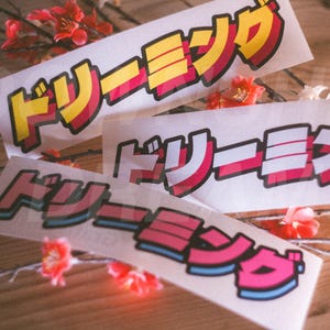 May include: Three rectangular stickers featuring the Japanese katakana text ドリーミング, which translates to dreaming. The stickers display bold, stylised lettering in yellow, pink, and blue with black outlines, arranged on a white background.