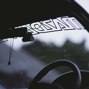 May include: A black car interior with a steering wheel and a rearview mirror. The windshield has a sticker that says "NZDR".