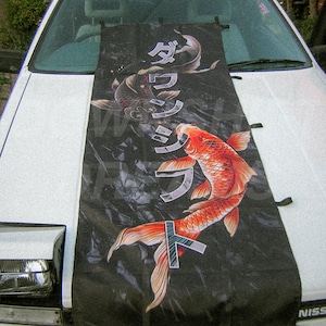May include: A black rectangular car hood cover featuring a graphic print of two koi fish, one orange and one dark grey, swimming in a stylized water pattern. The design includes vertical Japanese katakana text in white, layered over the fish imagery.