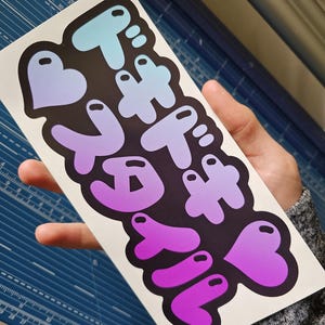 May include: A sticker featuring Japanese text in a gradient of blue, purple, and pink. The text is in a bubble-like font, set against a black background. The sticker is held in a person's hand.
