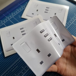 May include: Three white plastic electrical outlet covers with two USB ports each. The covers are designed to look like a standard UK three-pin plug socket.