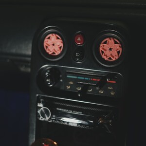 May include: Close-up of a black car dashboard with two pink flower-shaped air fresheners. The dashboard features a radio, climate control buttons, and a hazard light button.