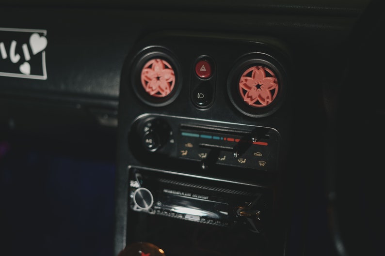 May include: Close-up of a black car dashboard with two pink flower-shaped air fresheners. The dashboard features a radio, climate control buttons, and a hazard light button.