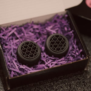 May include: Two black ear gauges with a heart pattern design, displayed in a black box with purple shredded paper.